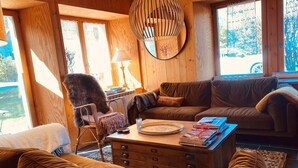 Interior acogedor de una cabaña, con paredes de madera, un sofá, una silla, una mesa con lámpara y una ventana con vistas al exterior.