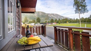 A wooden deck with a table holding a fruit bowl, a bottle, and a wine glass. A mountain and a road are visible in the background. - Topcamp Mosjøen - Helgeland (Vefsn)