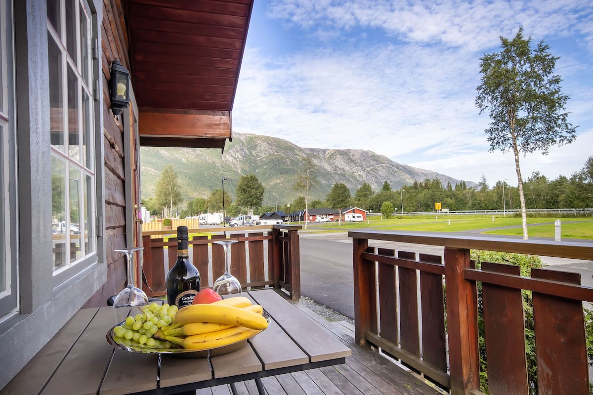 Una terraza de madera con una mesa que sostiene un frutero, una botella y una copa de vino. Se ven una montaña y una carretera al fondo.