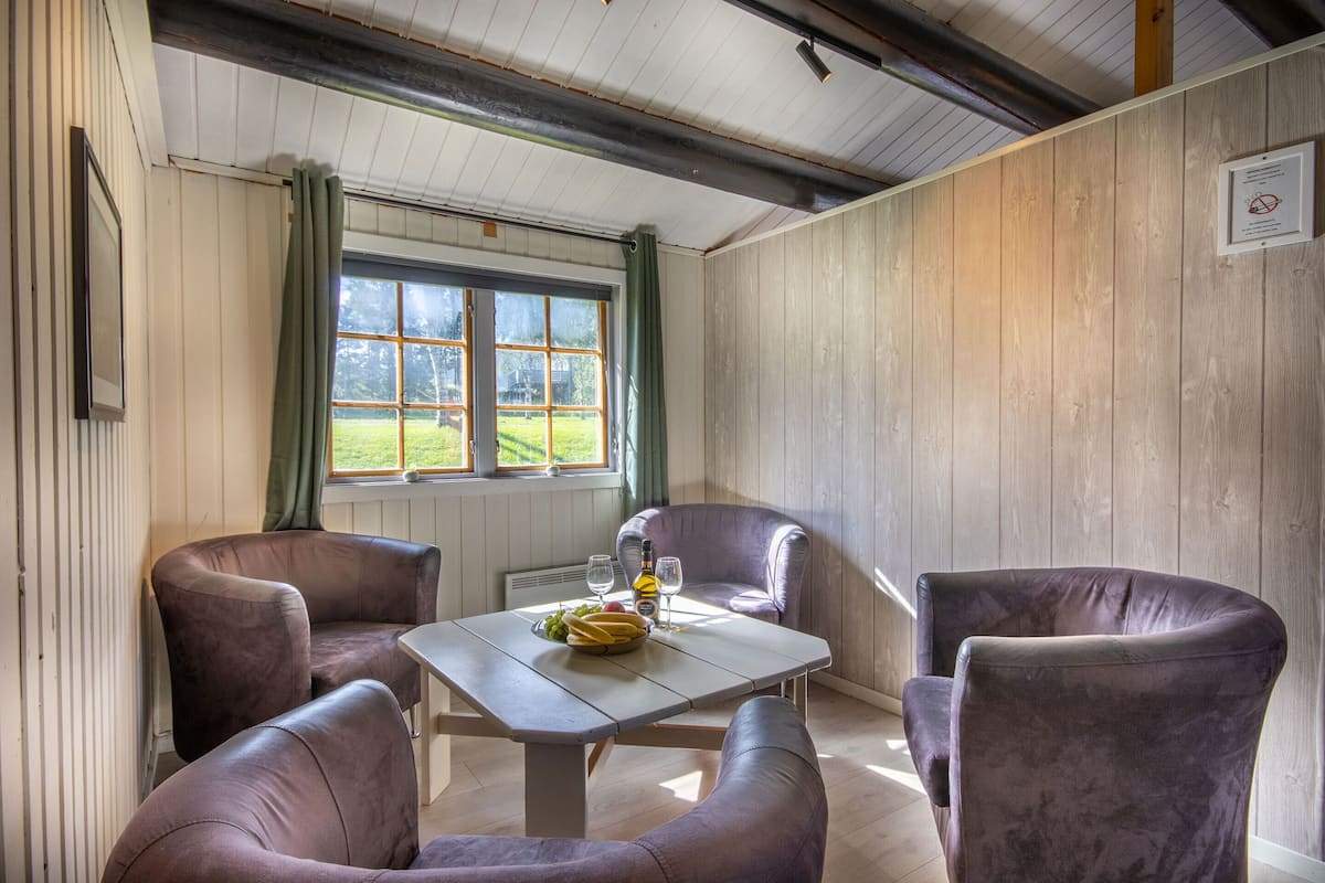 Una habitación con techo de madera, una ventana con cortinas, una mesa con una botella y copas, y dos sillas moradas.