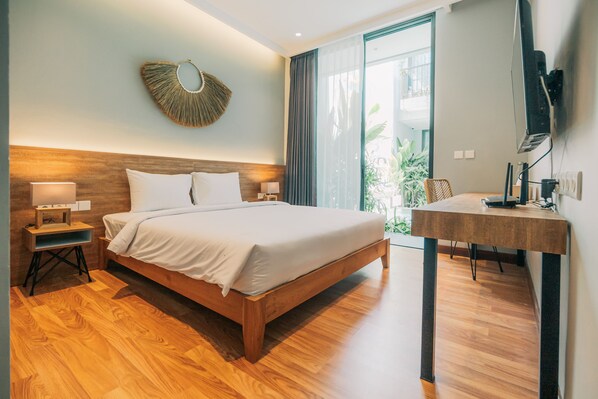 A modern hotel room with a wooden bed, a bedside table with a lamp, a wall-mounted TV, and a large window with a view of greenery.