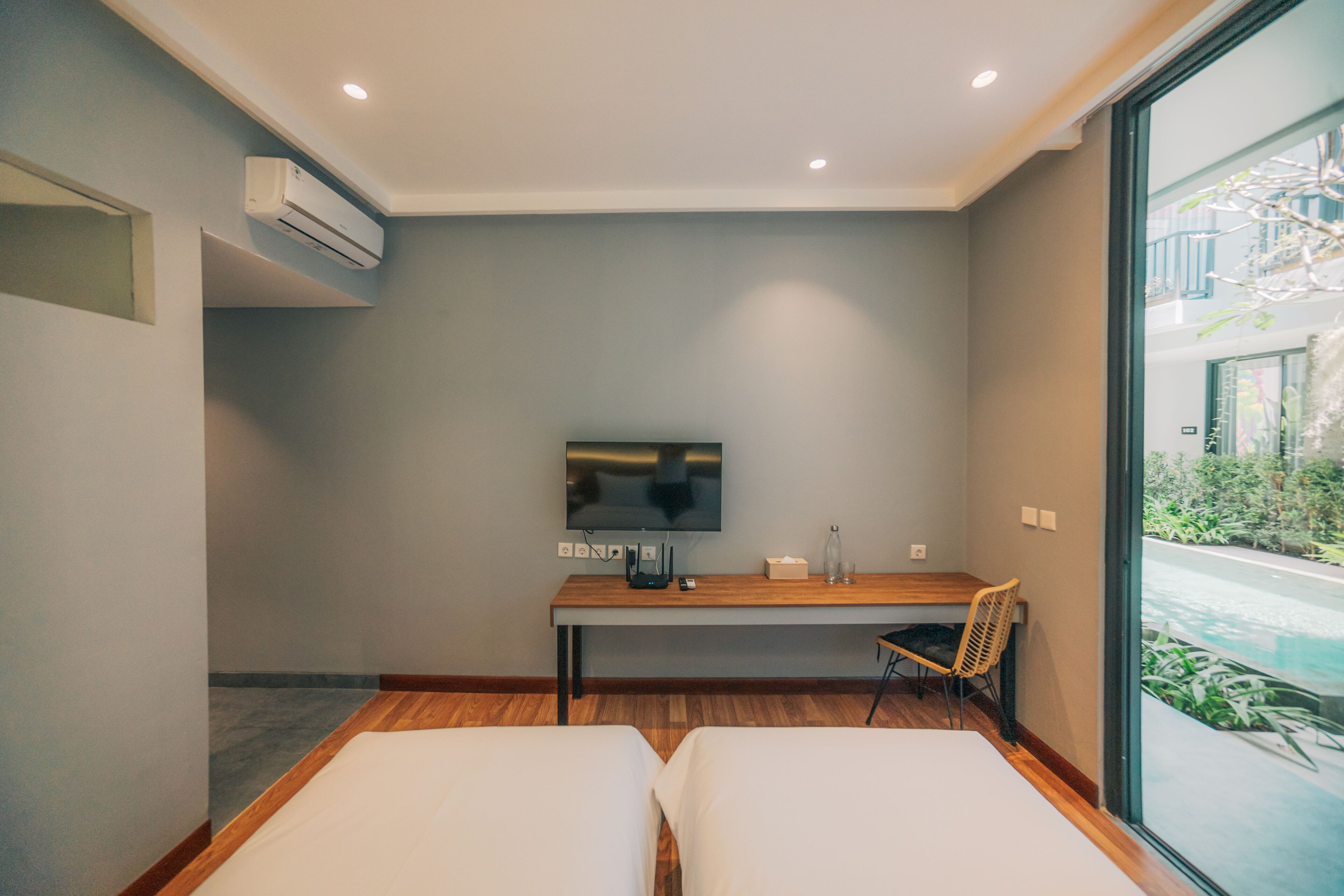 Une chambre d’hôtel moderne équipée d’un téléviseur à écran plat, d’un bureau en bois, de deux lits et d’une grande fenêtre donnant sur une piscine.