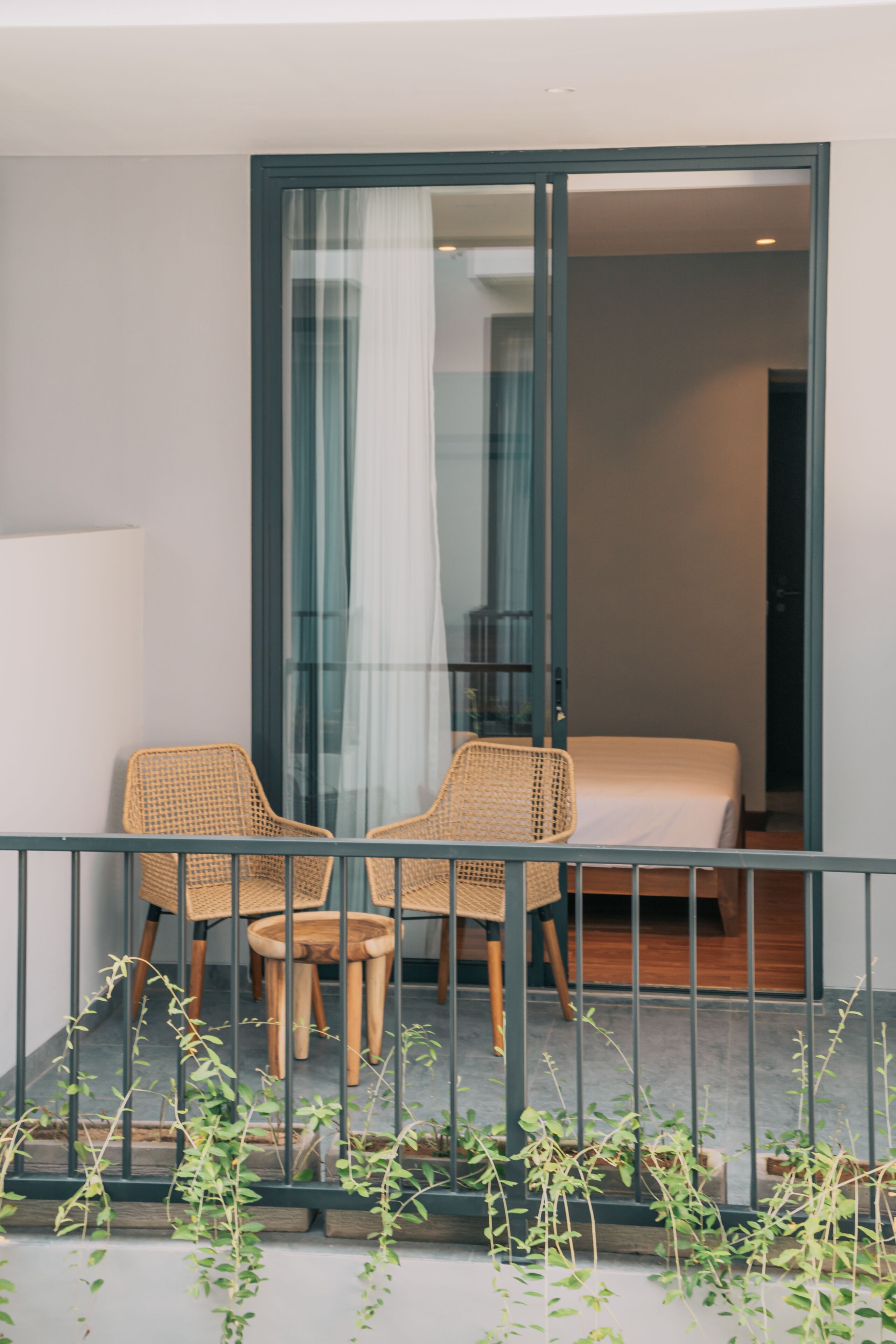 Een balkon met twee rieten stoelen en een tafeltje, een schuifdeur naar een slaapkamer met een bed, en uitzicht op de omgeving.