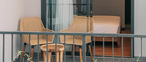 A balcony with two wicker chairs and a small table, a sliding glass door leading to a bedroom with a bed, and a view of the outside.