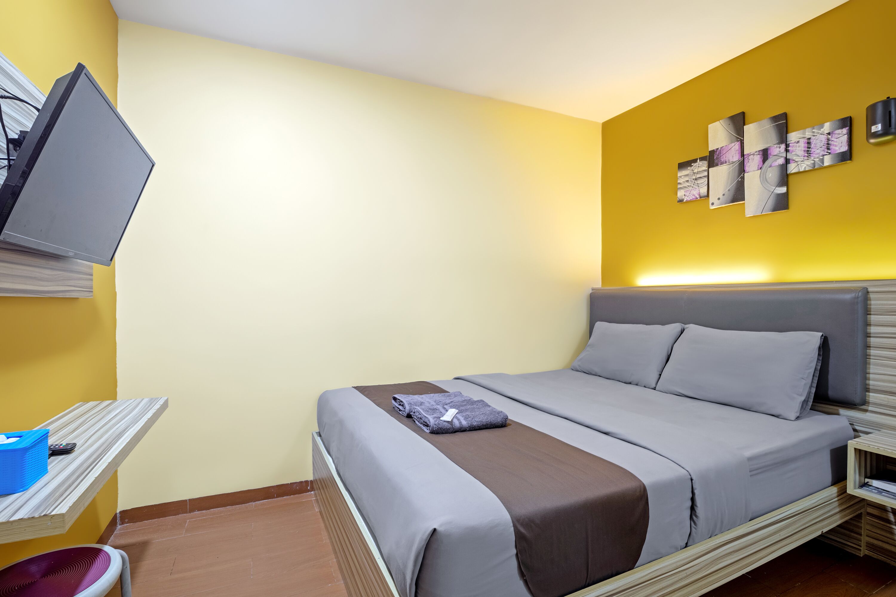 Een opgemaakt bed met grijs en bruin beddengoed, een televisie aan de muur en een klein tafeltje met een blauwe mand en afstandsbediening.