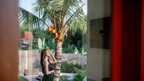 A woman in a green dress standing by a palm tree with coconuts, in a courtyard with a pool and greenery. - Musa Canggu by Zuzu (Tibubeneng)