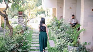 A pathway in a garden area with a person walking away from the camera. There are potted plants and a building with columns in the background. - Musa Canggu by Zuzu (Tibubeneng)