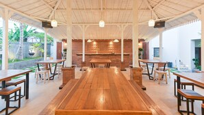 A spacious indoor dining area with wooden tables and benches, a brick wall with decorative elements, and a ceiling with exposed beams and hanging lights. - Musa Canggu by Zuzu (Tibubeneng)