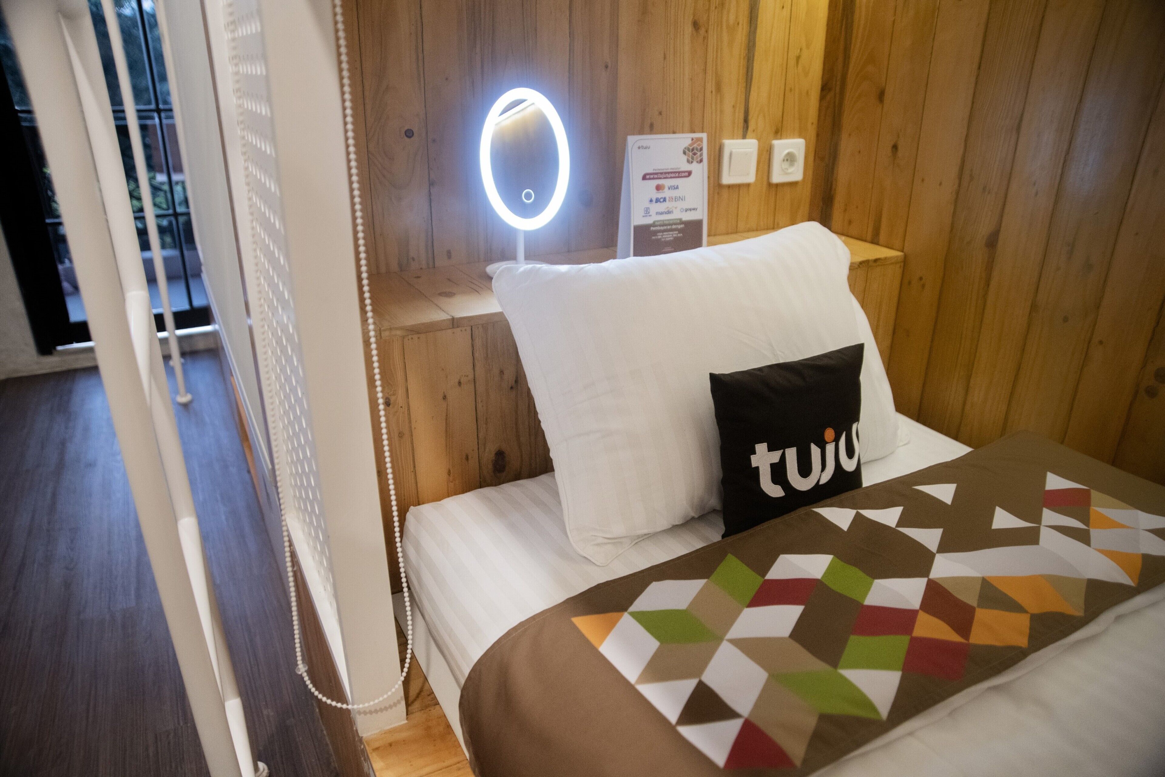 A hotel room with a bed, a bedside table with a ring light, a sign indicating payment options, and wooden paneling on the walls.