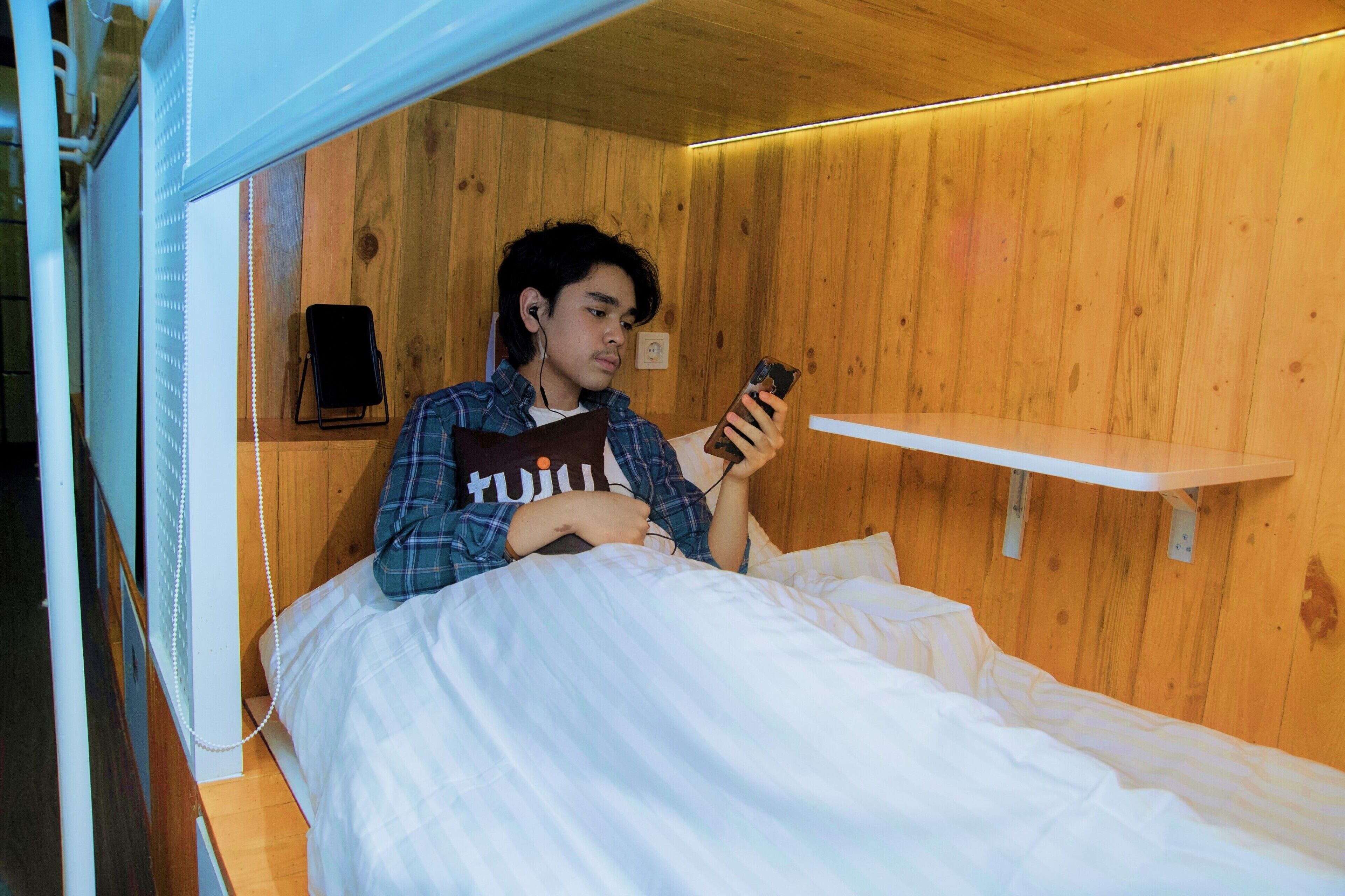 A person lying in a wooden cabin-style room with a bed, a white shelf, and a wall-mounted speaker.