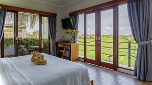 A bedroom with a bed, a desk, a TV, and a view of a green landscape through large sliding doors. - Canggu Hype (Canggu)