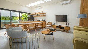A modern living room with a large TV, comfortable seating, a kitchen area, and a view of greenery outside. - Maitreya Villa (Jimbaran)