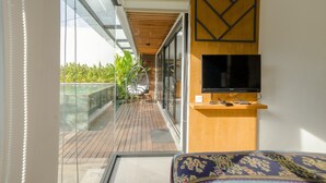 A balcony with a wooden floor, a glass railing, and a view of greenery. - Maitreya Villa (Jimbaran)