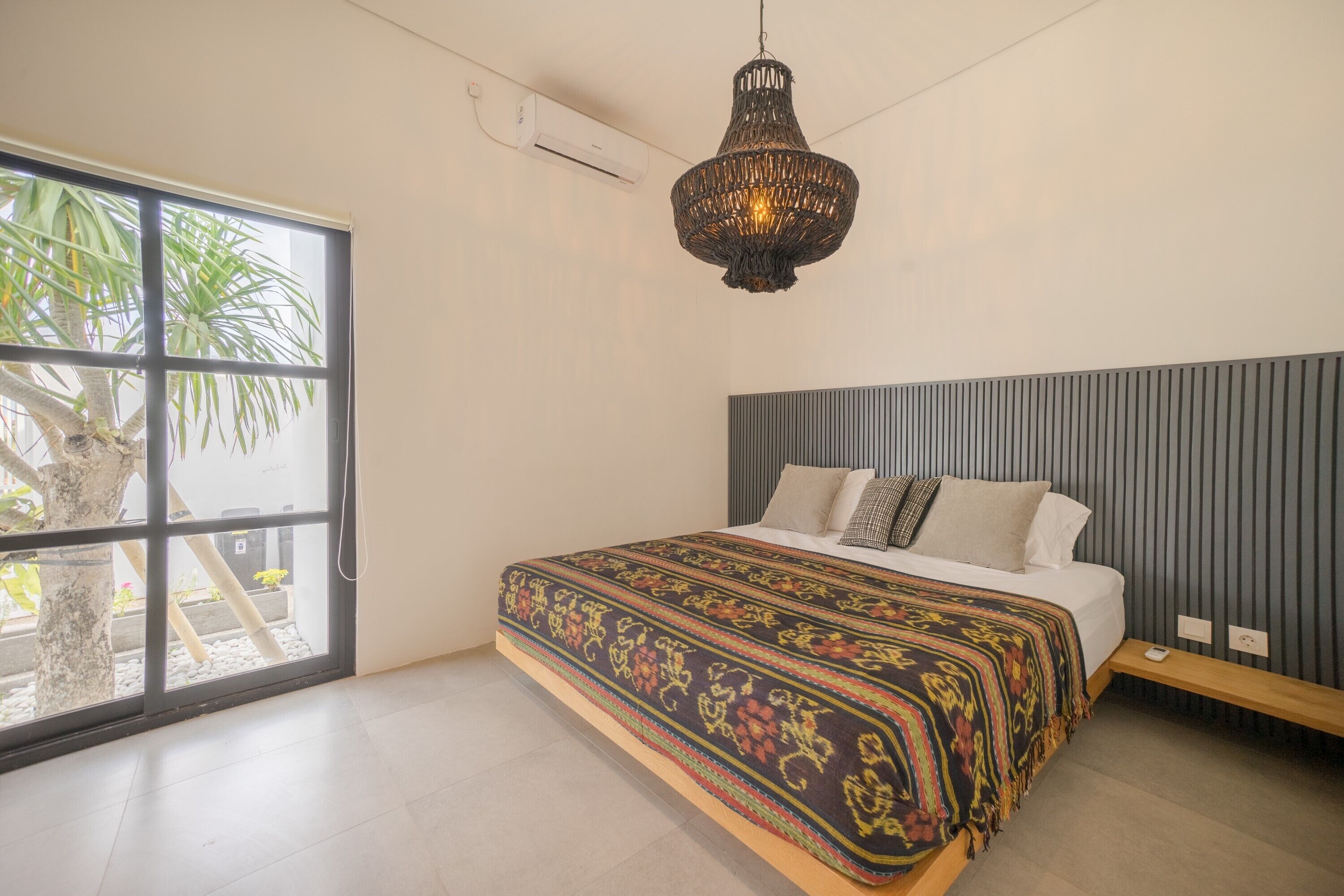 A modern bedroom with a large bed, a hanging light fixture, and a view of a tree outside.