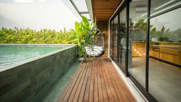 A wooden deck with a hanging chair, a pool, and a glass-enclosed area with plants. - Maitreya Villa (Jimbaran)