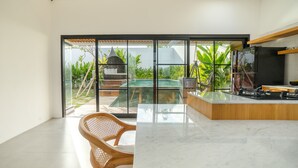 A modern kitchen with a marble countertop, wooden chairs, and a view of a pool and garden through large glass doors. - Maitreya Villa (Jimbaran)