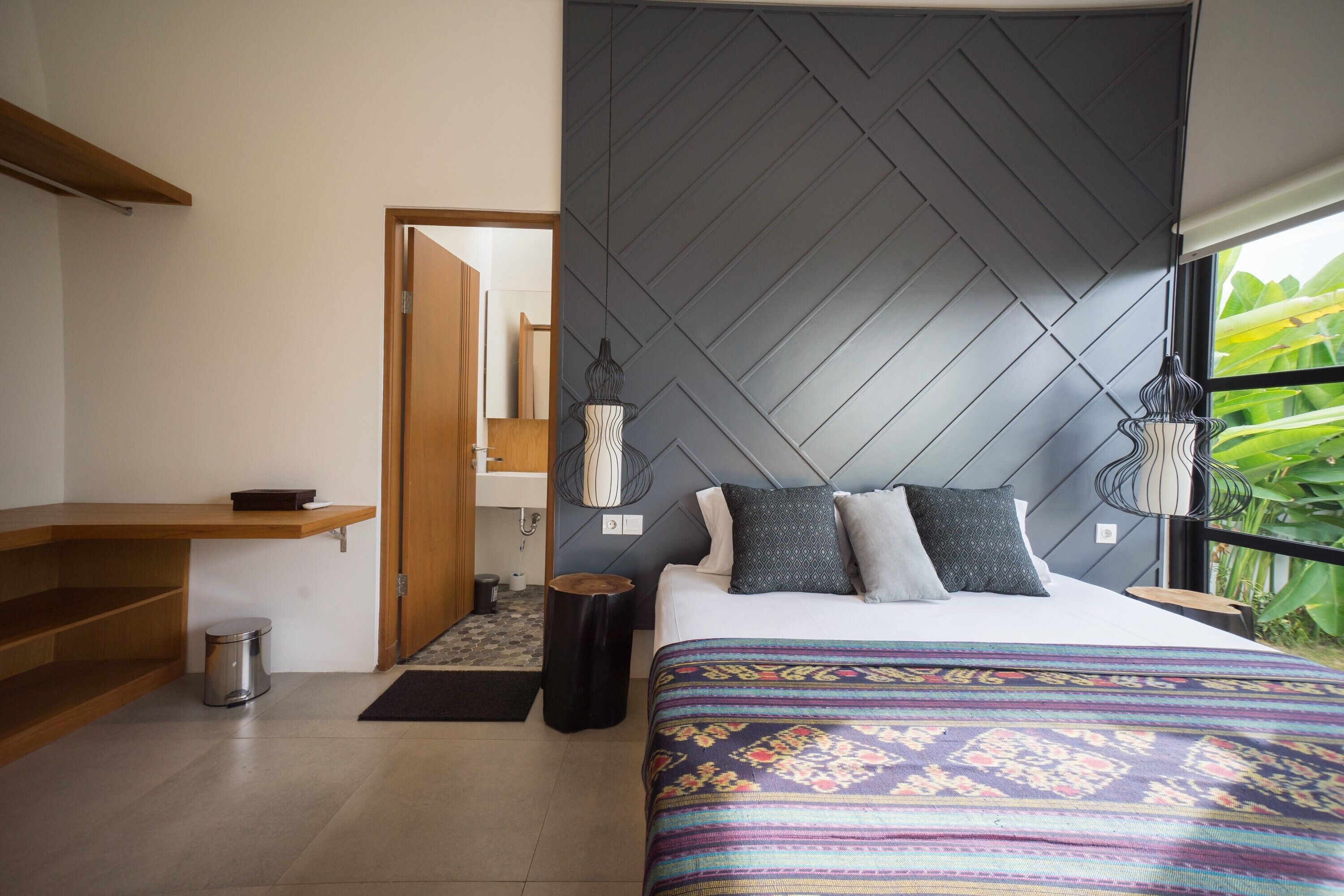A modern bedroom with a herringbone-patterned headboard, a large bed with patterned bedding, a wooden shelf, a small trash bin, and a window with a view of greenery.