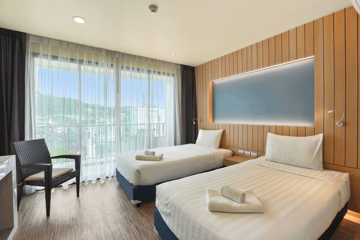 Habitación de hotel con dos camas, un escritorio, una silla y un gran ventanal con cortinas.