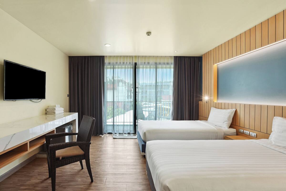 Habitación de hotel con televisión de pantalla plana, un escritorio, dos camas, una silla y un ventanal con cortinas.