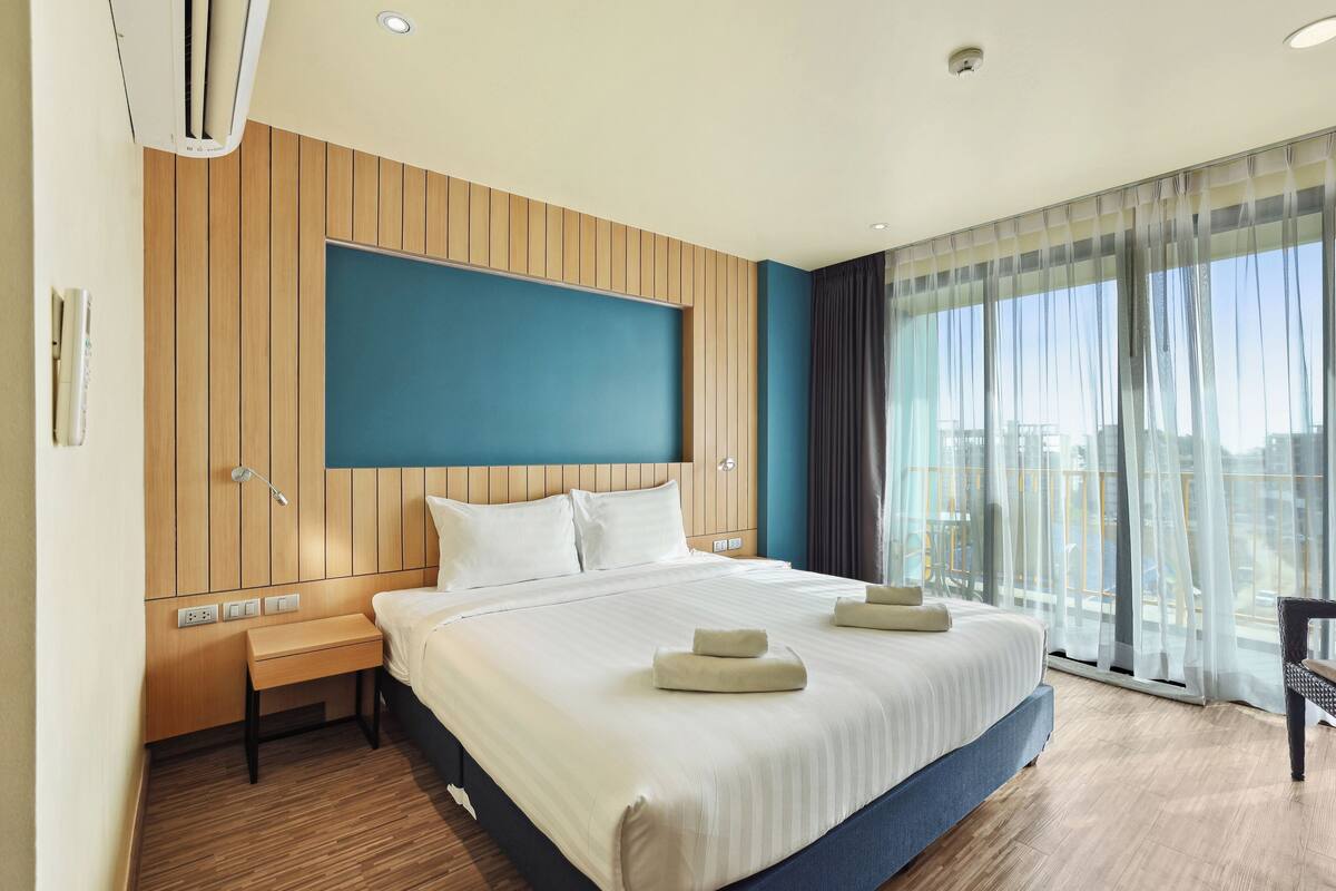 Habitación de hotel con una cama grande, paneles de madera en las paredes, una pared de acento azul, una mesita de noche y vistas de la ciudad a través de amplios ventanales.