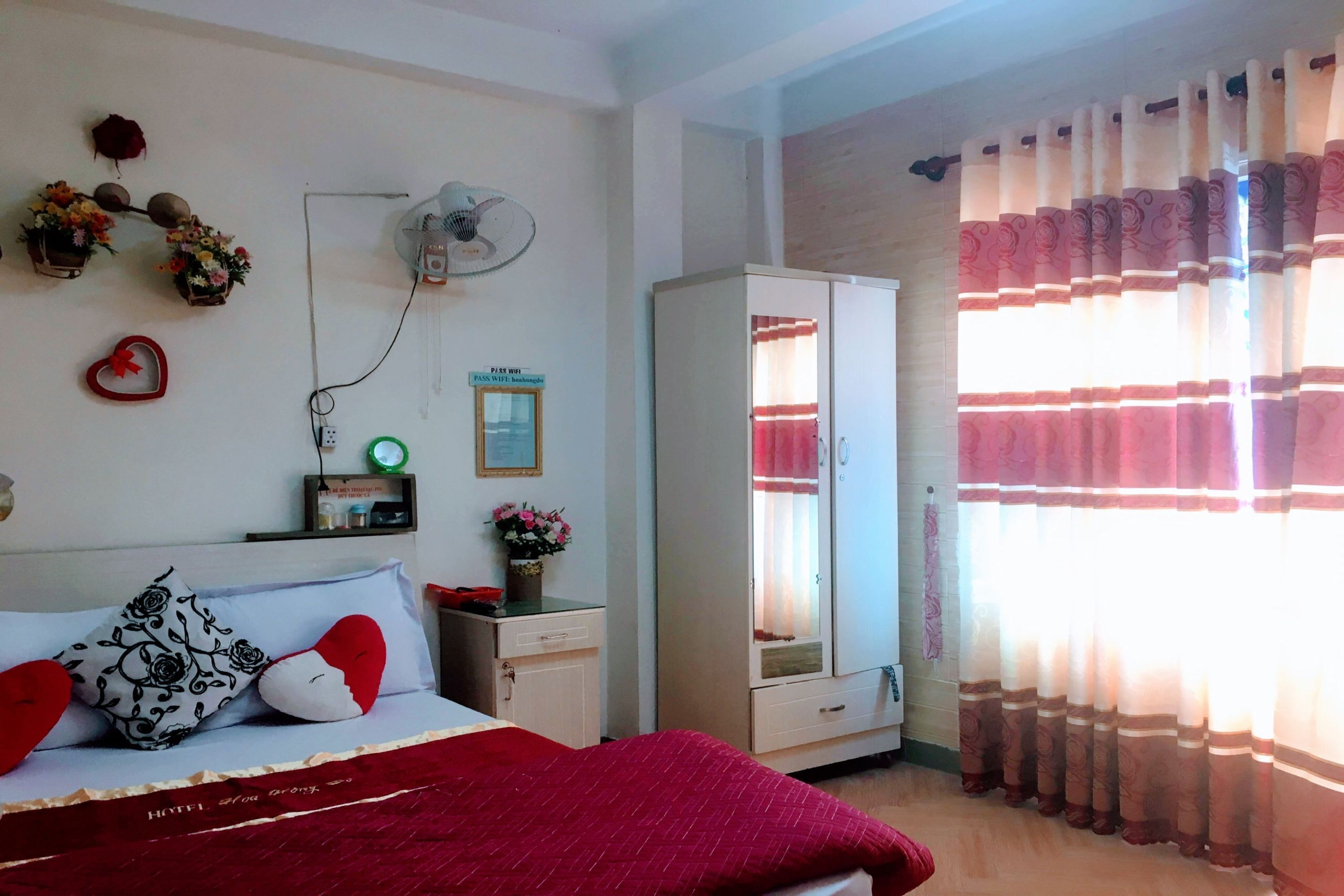 Quarto de hotel com cama, guarda-roupa, mesa de cabeceira com luminária, ventilador e janela com cortinas.