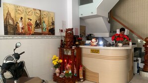 Taulell de recepció amb un cartell que diu "QUÁY LỆ TÂN RECEPTION", una moto aparcada a l'esquerra i una paret amb un rellotge i una pintura d'una dona amb vestimentes tradicionals.