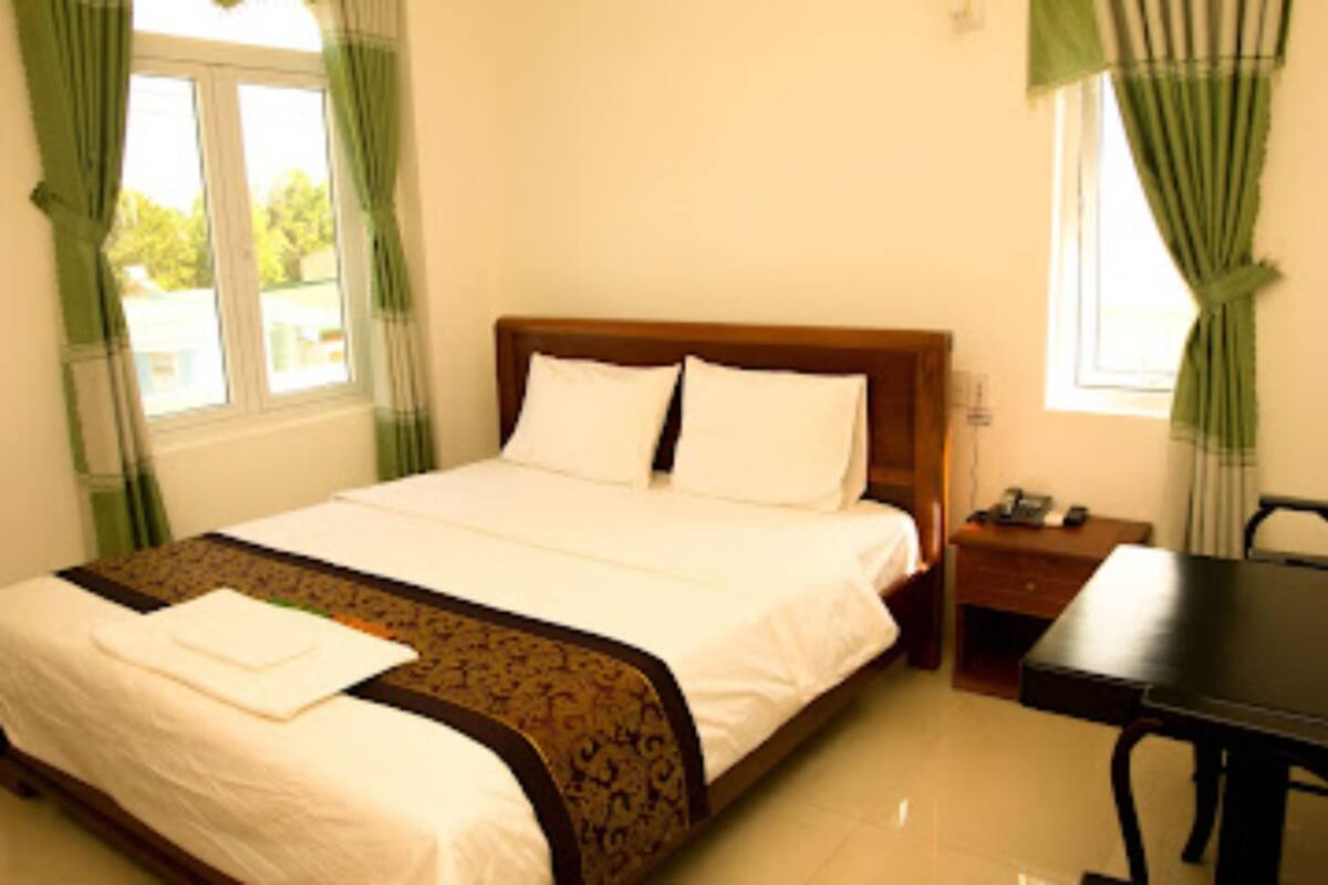 A hotel room with a bed, a wooden nightstand, a desk, and a window with curtains.