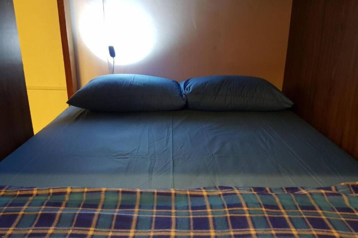 A bed with a blue mattress and pillows, a plaid blanket, and a wall-mounted light fixture.