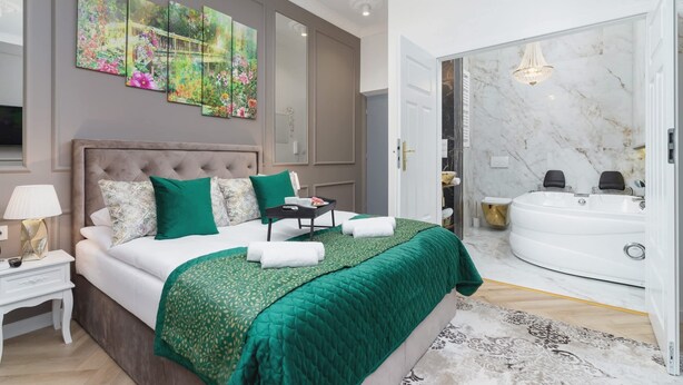 A bedroom with a large bed, green bedding, a nightstand, a bedside lamp, a painting, a bathroom with a marble wall, a bathtub, and a chandelier.