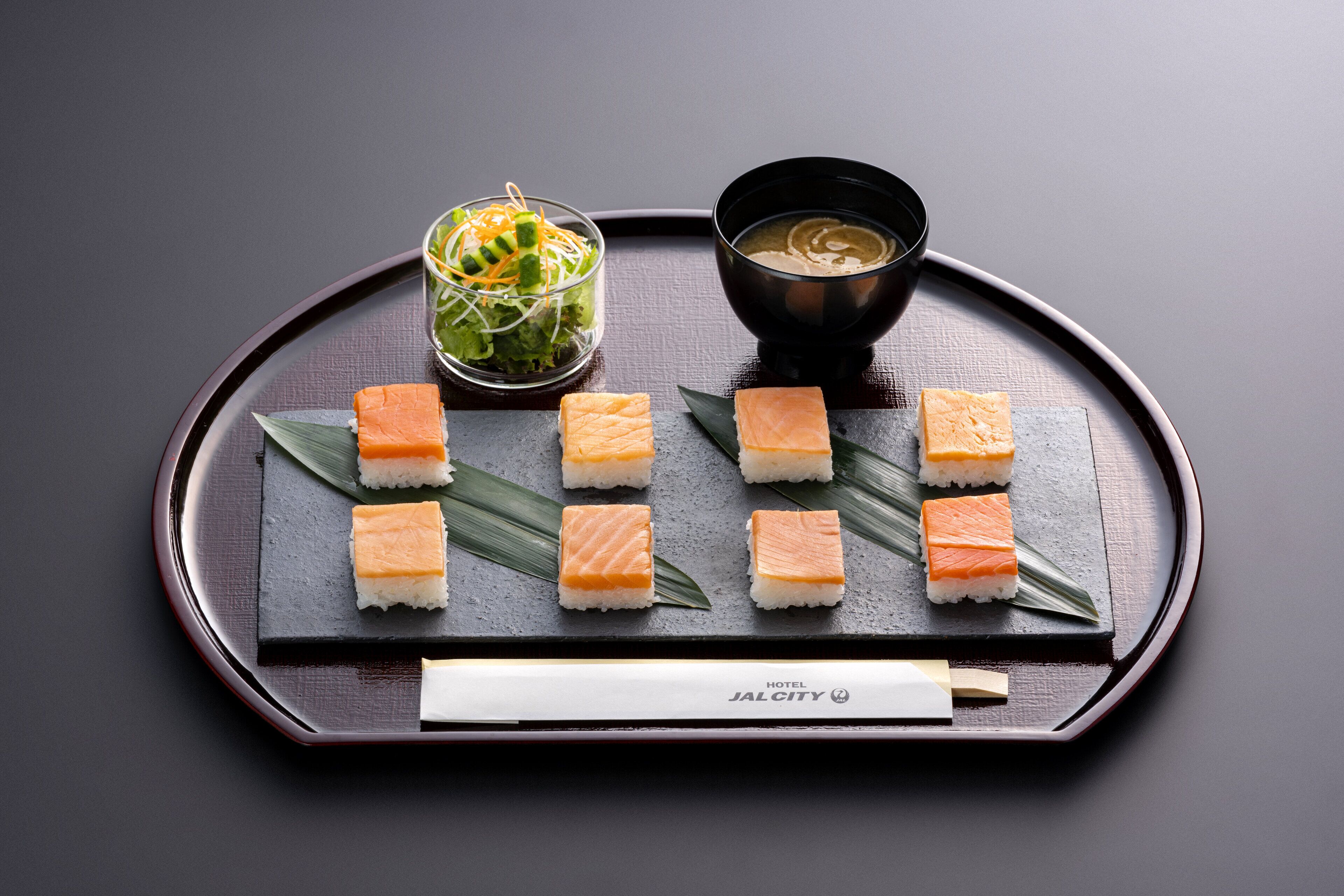 Una bandeja con sushi, una ensalada verde en un vaso y un tazón de sopa.