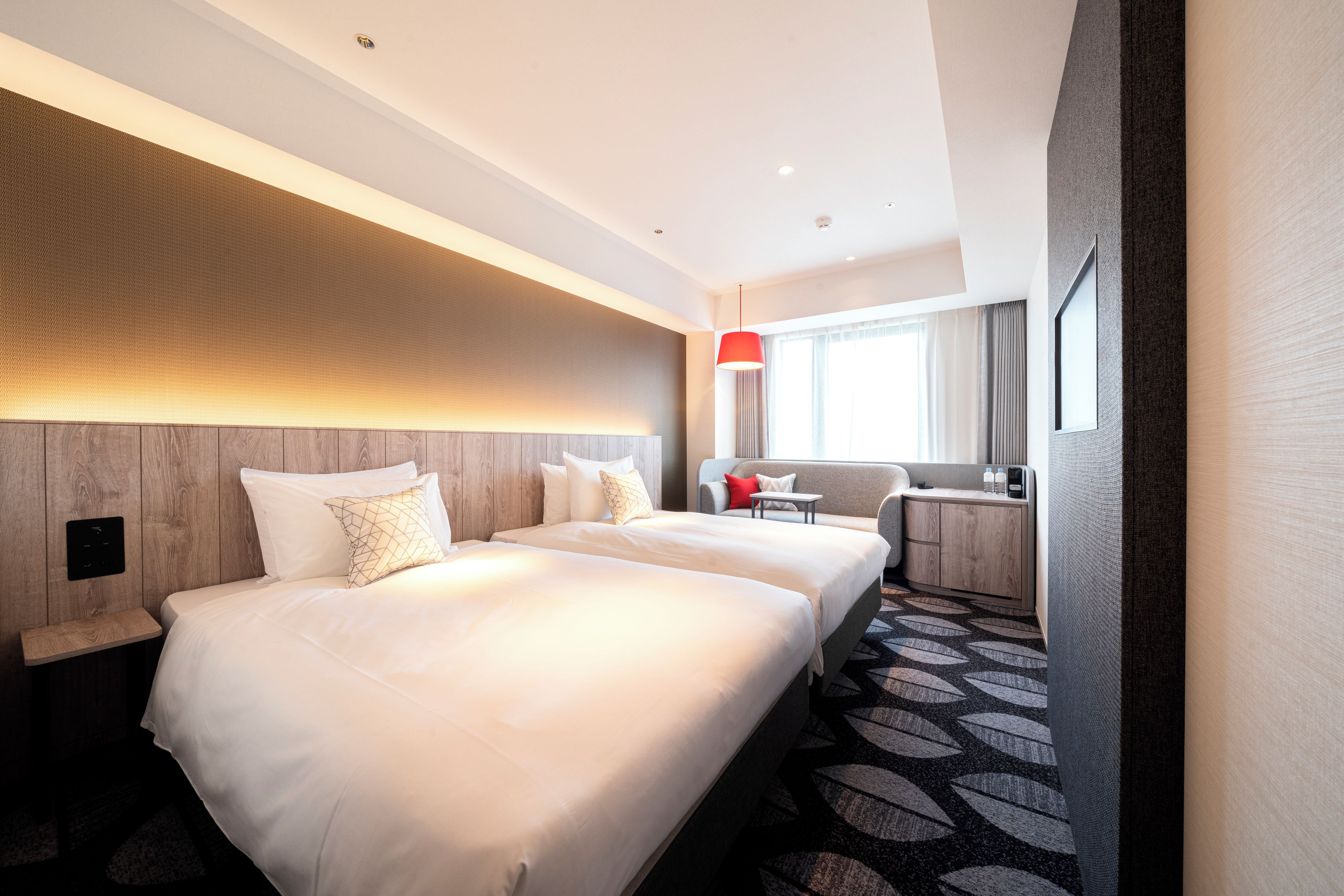 Ein Hotelzimmer mit zwei Betten, einem Schreibtisch, einem Stuhl, einer roten Pendelleuchte und einem großen Fenster mit Vorhängen.