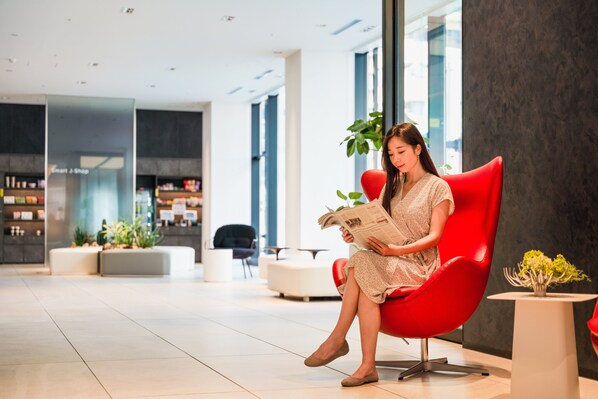 Una mujer sentada en una silla roja en un vestíbulo moderno con una planta, una mesa blanca y una Smart J-Shop al fondo.