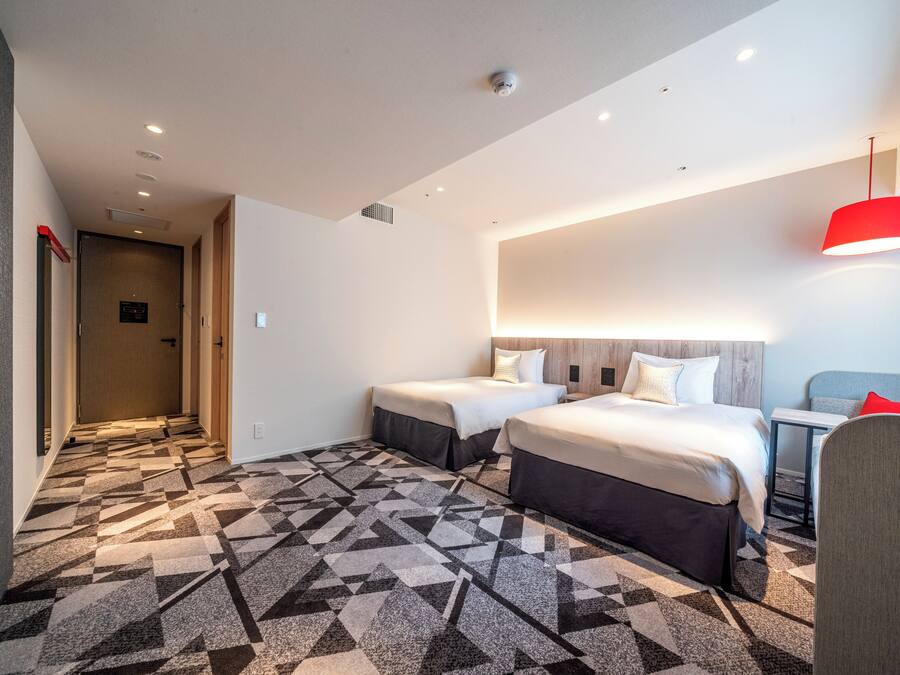 A hotel room with two beds, a patterned carpet, a red pendant light, and a hallway with a door.