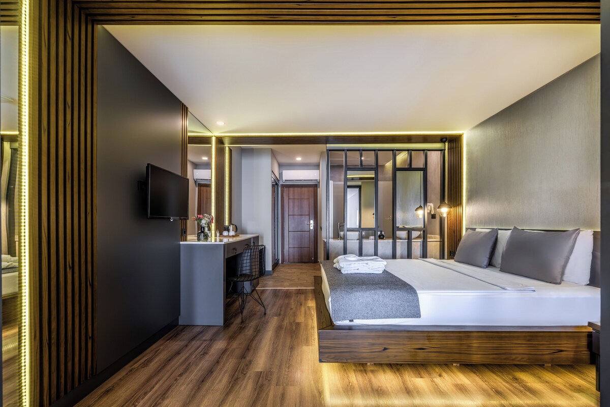 A modern hotel room with a large bed, a desk with a flat-screen TV, and a bathroom visible through a sliding door.