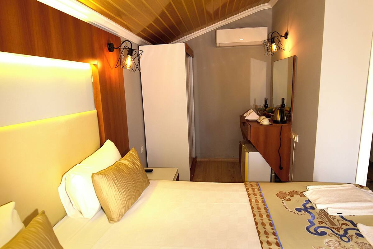 Habitación de hotel con techo de madera, cama con ropa blanca, mesita de noche, espejo y baño con lavamanos y espejo.