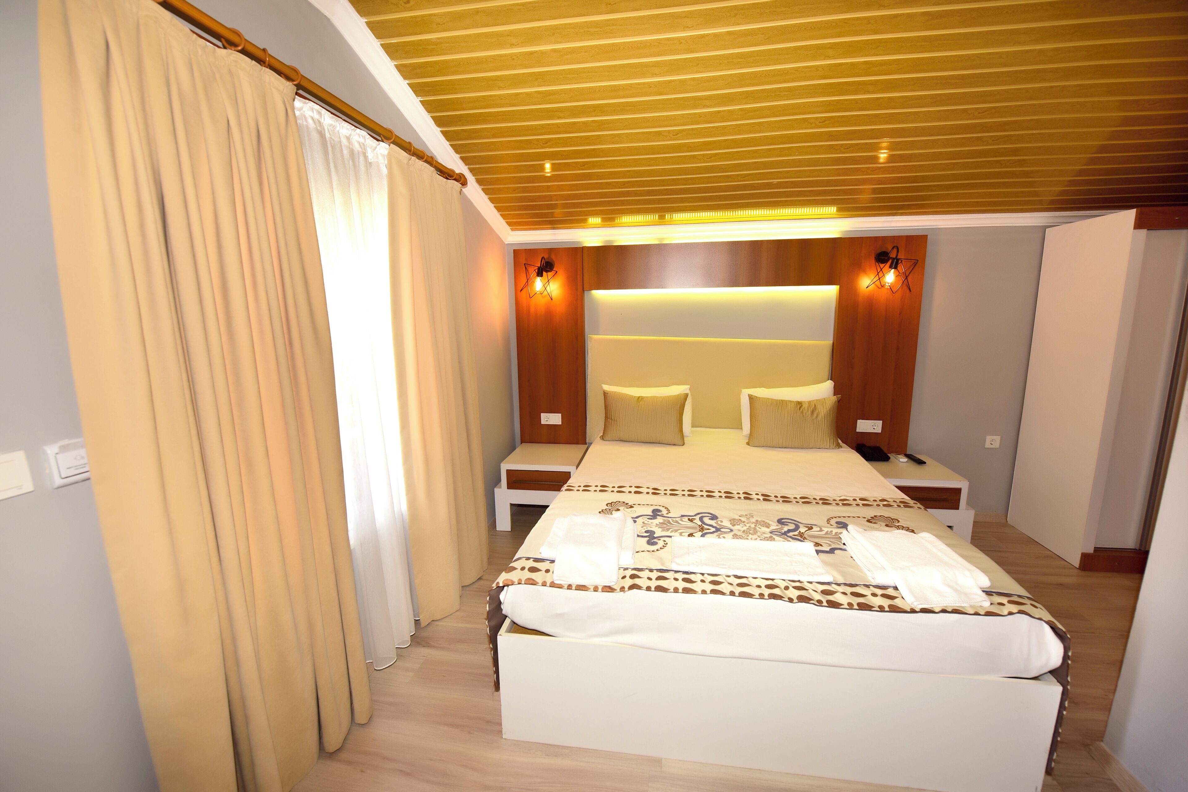 A hotel room with a large bed, bedside tables, a wooden ceiling, and wall-mounted lighting.