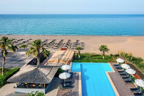 Un área de piscina con sillas de descanso y sombrillas, una playa con sombrillas y palmeras, y una vista despejada del océano.