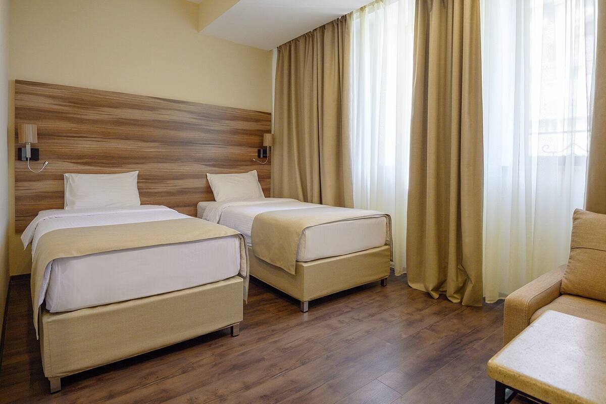 Habitación de hotel con dos camas, un sofá, cabecero de madera y ventanas grandes con cortinas.
