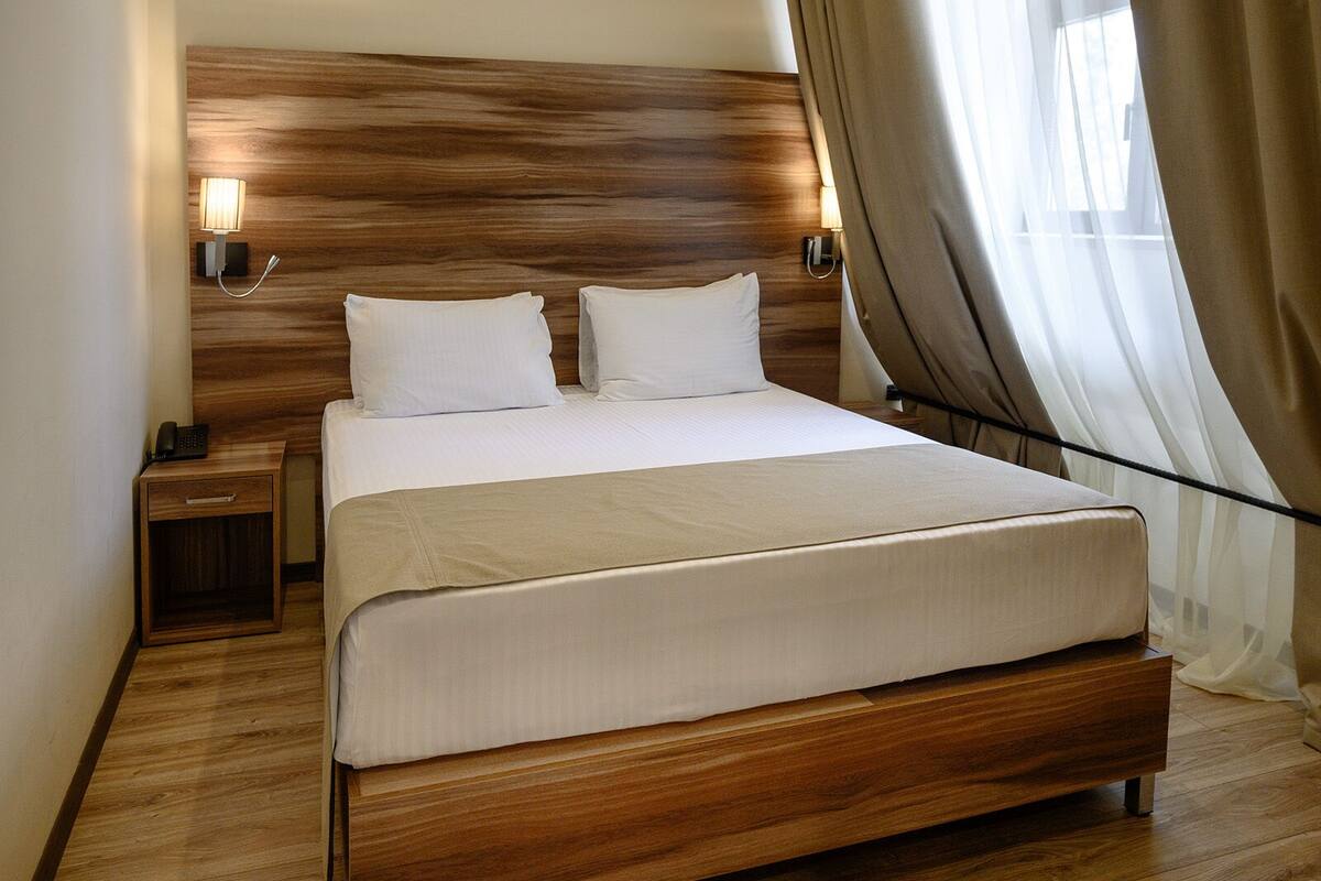 Una cama bien tendida con un cabecero de madera, dos lámparas de noche y un teléfono sobre una mesita de noche de madera.