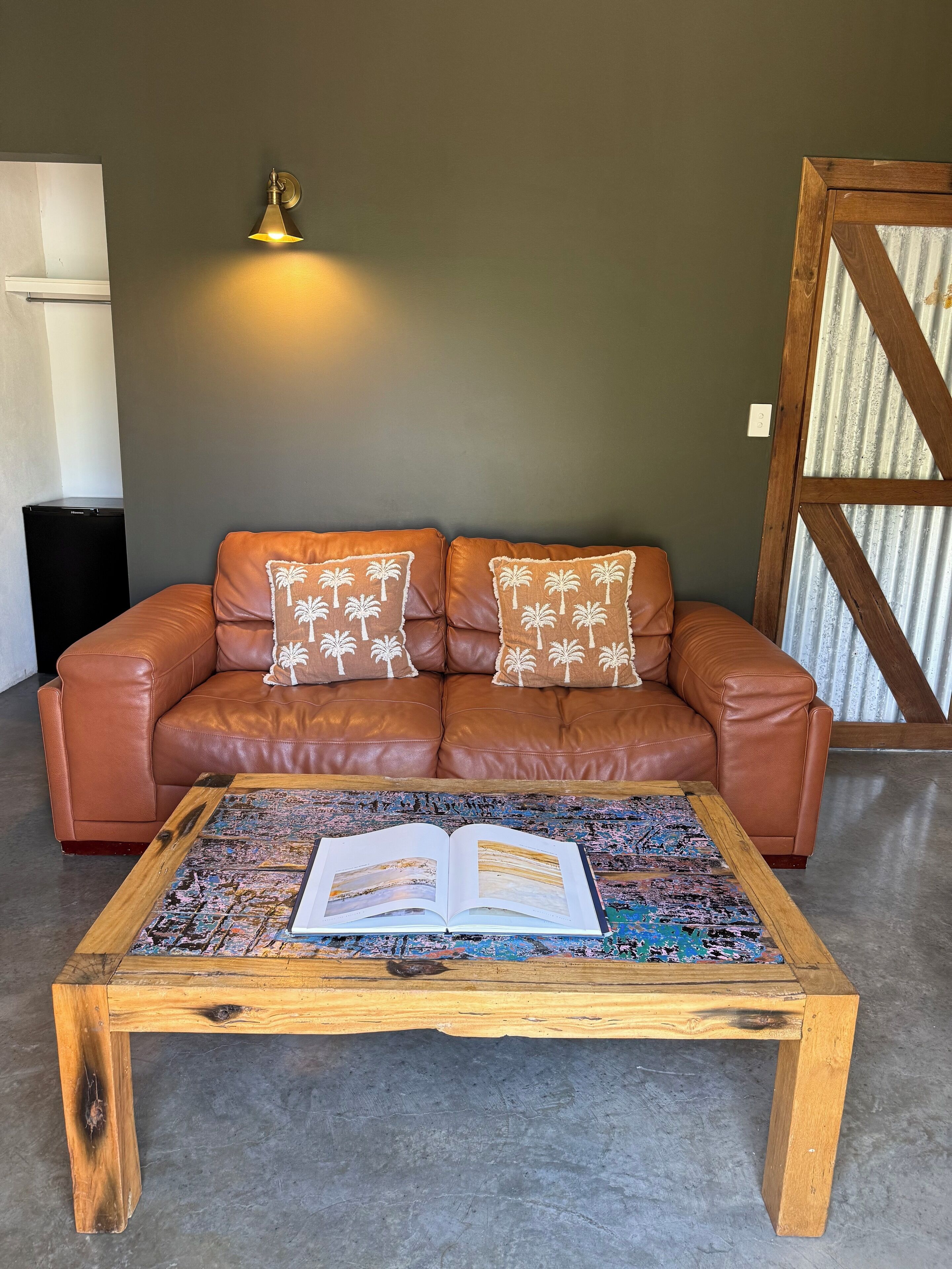 Un sofá de cuero marrón con dos almohadones decorativos frente a una pared verde, una mesa de centro de madera con un libro abierto, y una lámpara fijada a la pared.