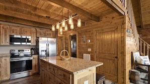 A wooden cabin-style kitchen with a central island, stainless steel appliances, and a hanging light fixture. - Upscale Cabin With Stunning Views (Boone)