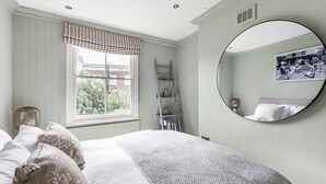 A bedroom with a bed, a window with a striped valance, a ladder, a shelf with books, and a round mirror on the wall.