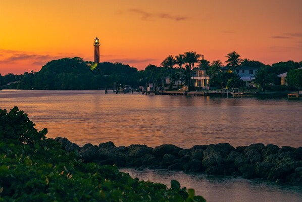 A coastal scene with a lighthouse, palm trees, and a calm sea at sunset.