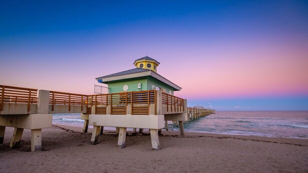 Una playa con un muelle de madera que conduce a un edificio verde con una torre, con vistas al océano.