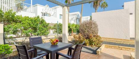 An outdoor patio with a table and chairs, a bowl of fruit, and a view of a white building with a balcony.