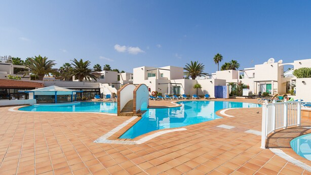 A pool area with tiled flooring, a clear blue sky, and white buildings with balconies. - Modern apartment. Swimming pool and beachfront. Corralejo (Corralejo)