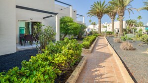 A modern building with a patio area featuring a tiled walkway, lush greenery, and palm trees. - Modern apartment. Swimming pool and beachfront. Corralejo (Corralejo)