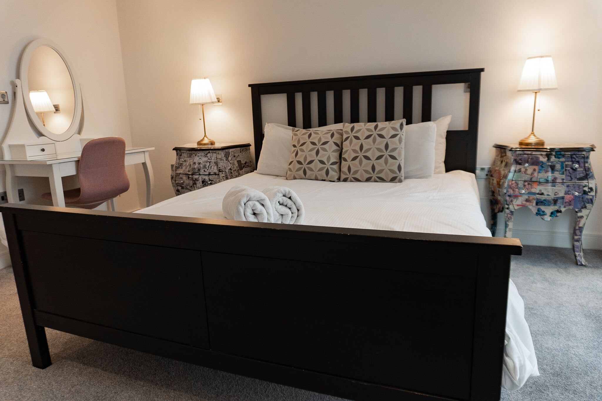 A neatly made bed with two towels, a bedside table with a lamp, a chair, and a dresser with a mirror.