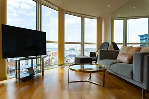 Uma sala de estar moderna com uma TV grande, um sofá, uma mesa de centro e grandes janelas com vista para a cidade.