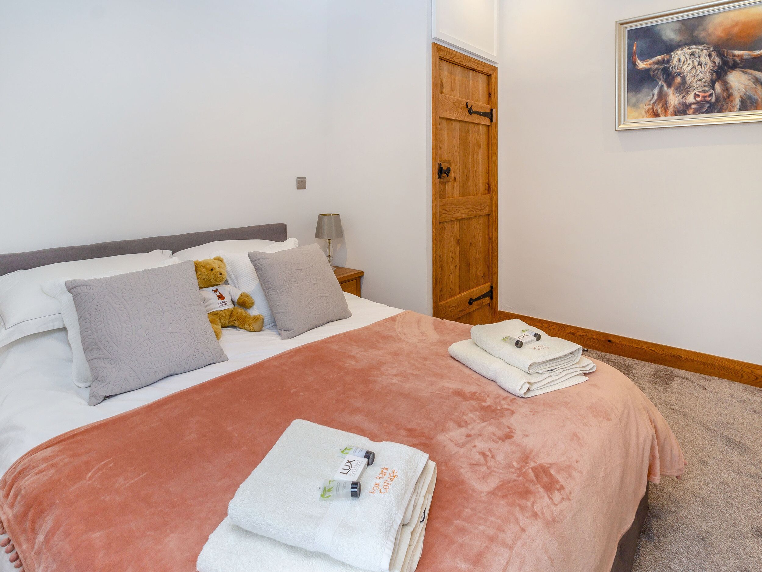 A neatly made bed with a pink blanket, pillows, and a teddy bear. There are towels and toiletries on the bed.